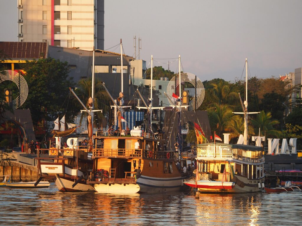pexels-photo-18230813-18230813-1 Rustic wooden sailboats docked at a vibrant city harbor during sunset, creating a picturesque urban scene.
