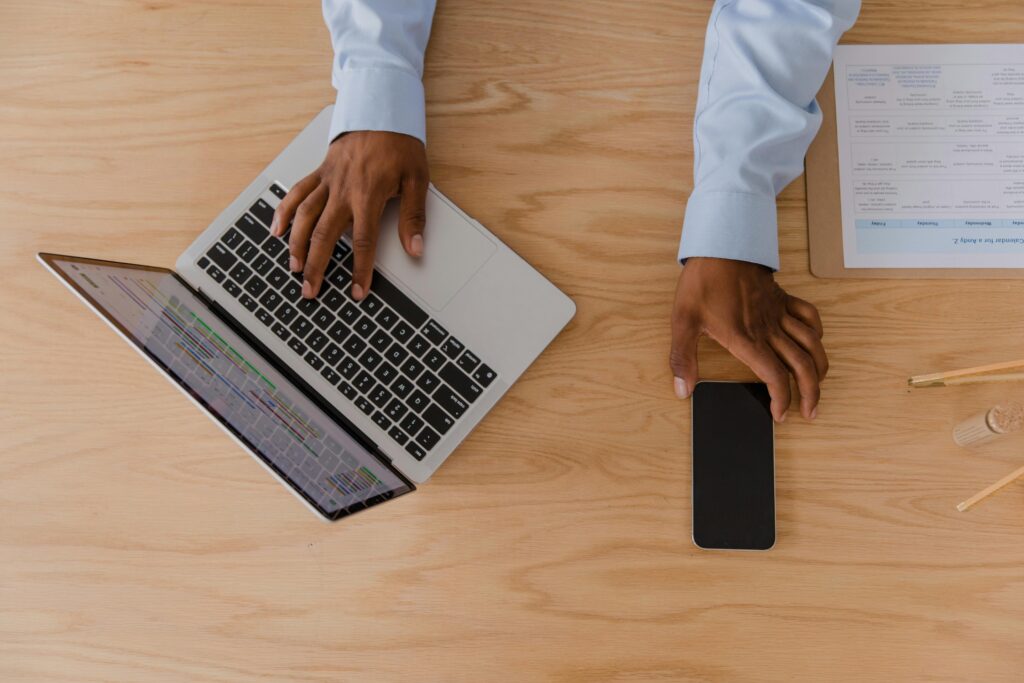 pexels-photo-10376213-10376213 Top view of a workspace featuring hands using a laptop and smartphone on a wooden desk.
